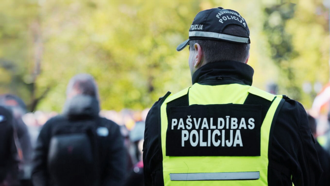ttēlā no mugurpuses redzams pašvaldības policijas darbinieks dzeltenā atstarojošā vestē ar uzrakstu „Pašvaldības policija”. Fonā redzami izplūduši cilvēki un zaļi koki.