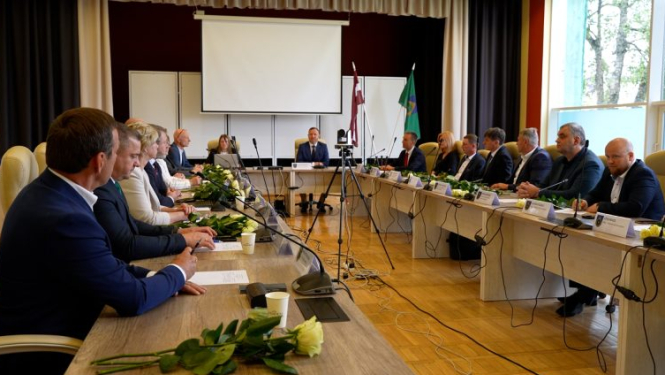 Talsu novada domes sēde zālē, kur deputāti un amatpersonas sēž pie galdiem abās pusēs, bet priekšā redzams runātājs un sēdes tehniskais aprīkojums.