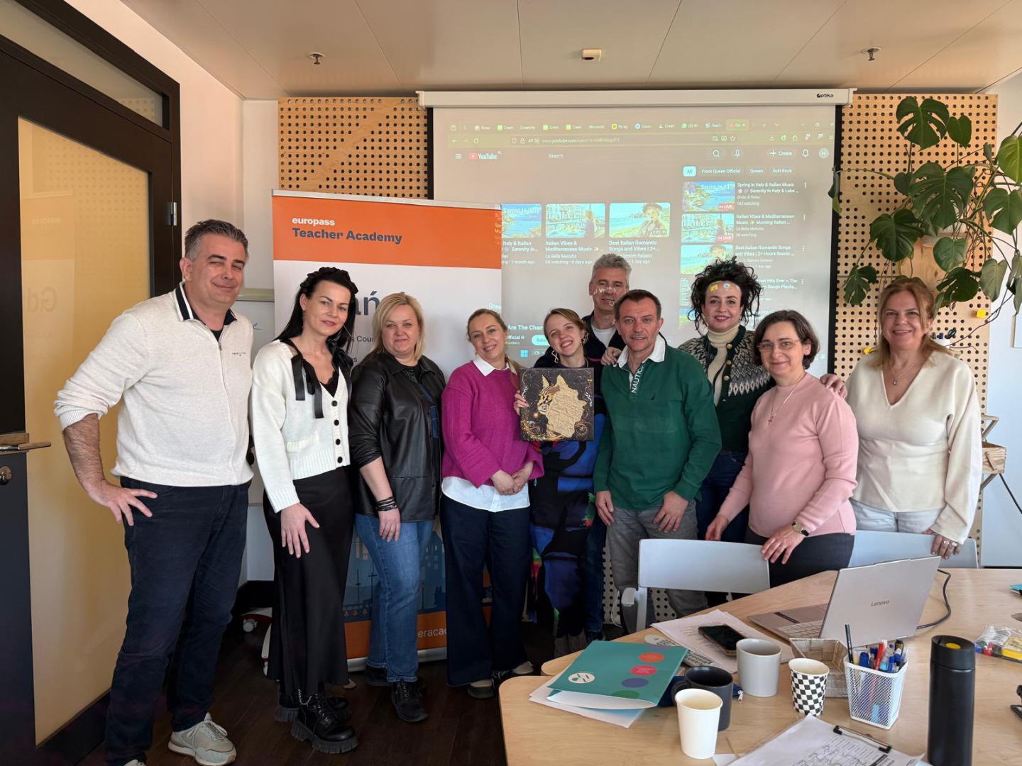 Grupas foto no Erasmus+ mobilitātes Gdaņskā – telpā pie „Europass Teacher Academy” stenda un projekcijas ekrāna pozē vairāki kursu dalībnieki. Priekšplānā redzams galds ar klēpjdatoru, krūzēm, rakstāmpiederumiem un mācību materiāliem, bet daļa dalībnieku rokās tur dekoratīvu darbu.