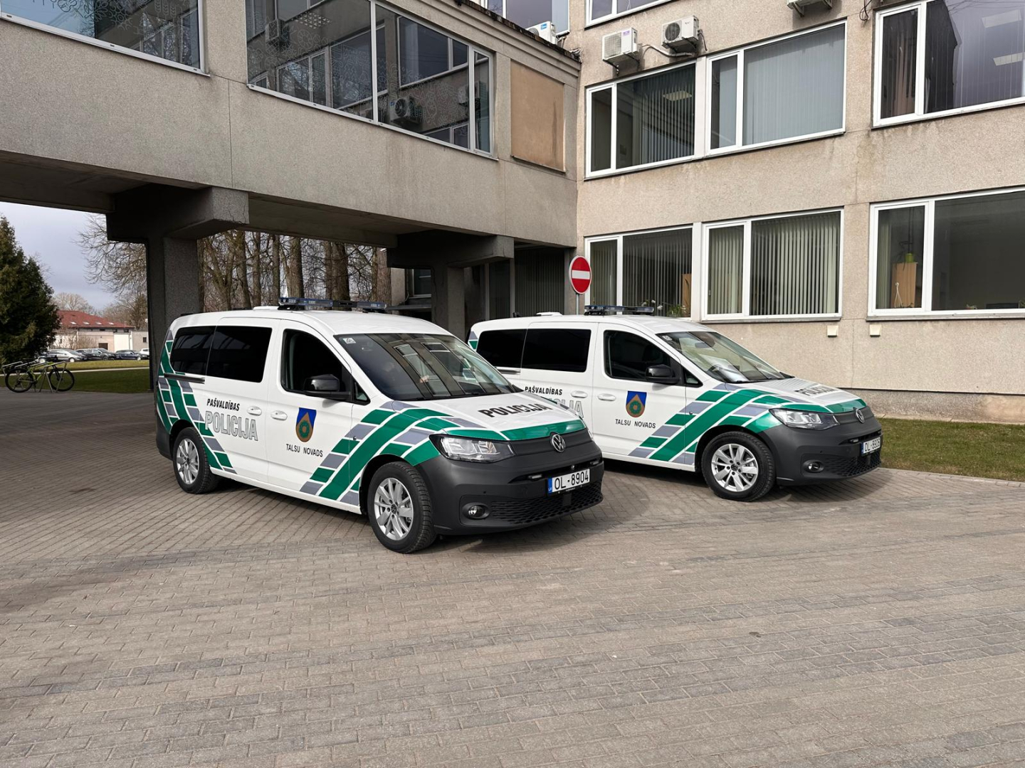 Attēlā pie administratīvās ēkas novietoti divi jauni Talsu novada Pašvaldības policijas operatīvie transportlīdzekļi – balti Volkswagen Caddy ar zaļi pelēku trafarējumu, uzrakstiem „Pašvaldības policija” un Talsu novada simboliku. Abiem transportlīdzekļiem uz jumta redzamas bākugunis.