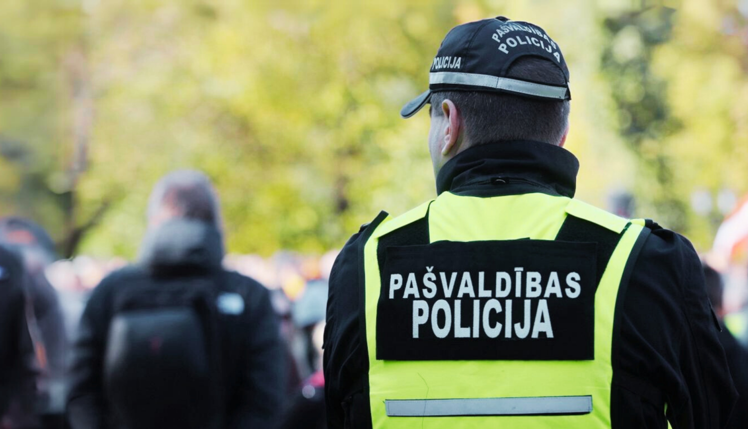 ttēlā no mugurpuses redzams pašvaldības policijas darbinieks dzeltenā atstarojošā vestē ar uzrakstu „Pašvaldības policija”. Fonā redzami izplūduši cilvēki un zaļi koki.