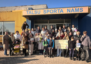 Pie ēkas ieejas redzama cilvēku grupa, kas svinīgi sagaida sportistu, turot rokās apsveikuma plakātu.