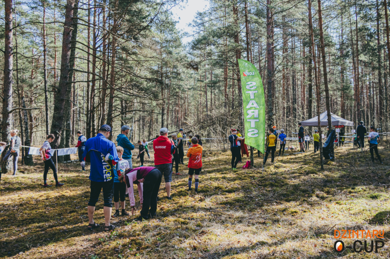 Orientēšanās sacensību centrs priežu mežā – dalībnieki un līdzjutēji pulcējušies pie starta zonas ar norobežojuma lentēm, redzams informatīvs karogs un telts.