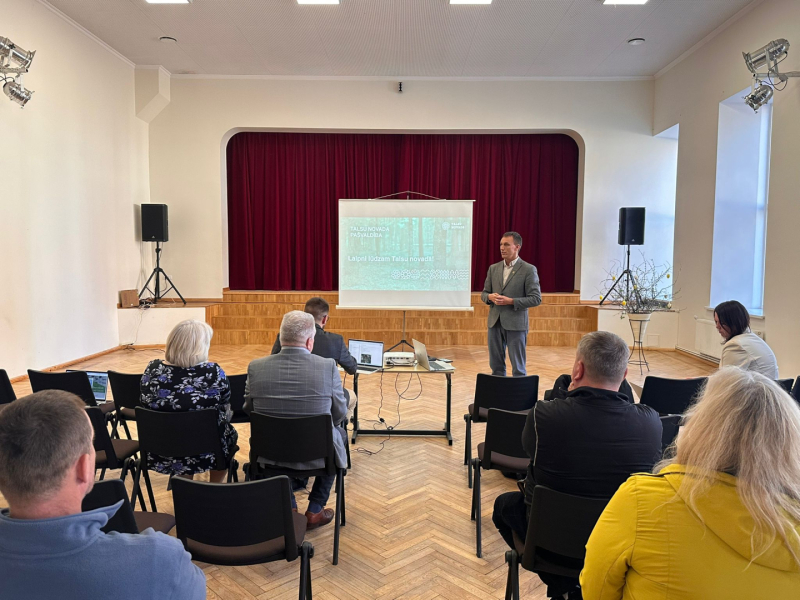 : Laucienes kultūras namā notiek Talsu novada pašvaldības tikšanās ar iedzīvotājiem. Zāles priekšā pie ekrāna stāv runātājs, bet klausītāji sēž krēslu rindās un seko prezentācijai. Fonā redzama skatuve ar tumši sarkaniem aizkariem, skaļruņi abās pusēs un galds ar projektoru un portatīvajiem datoriem.