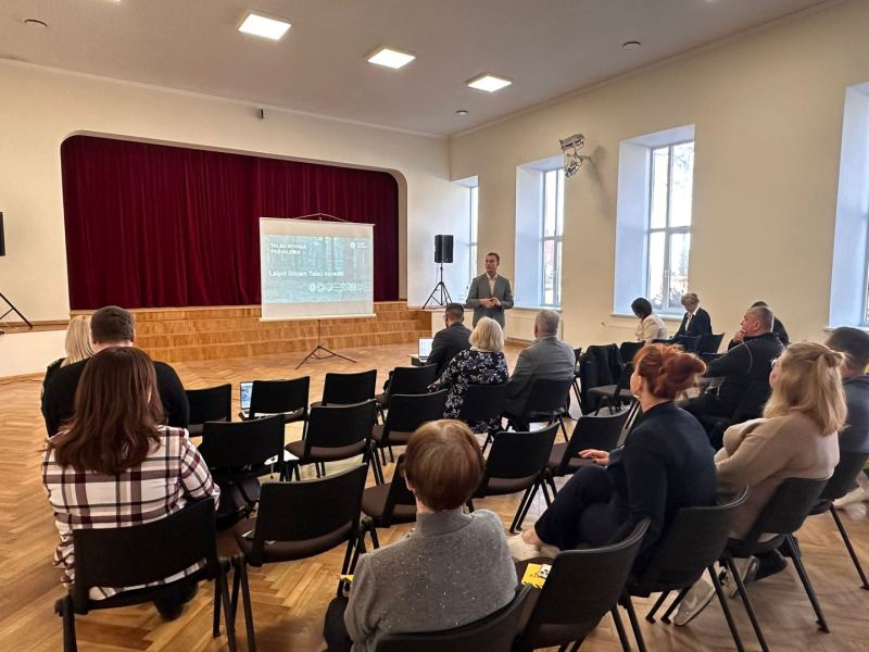 Laucienes kultūras nama zālē notiek Talsu novada pašvaldības tikšanās ar pagasta iedzīvotājiem. Zāles priekšā pie ekrāna stāv runātājs, bet iedzīvotāji sēž krēslos un klausās prezentāciju. Fonā redzama skatuve ar tumši sarkaniem aizkariem, bet labajā pusē – augsti logi, kas izgaismo telpu.