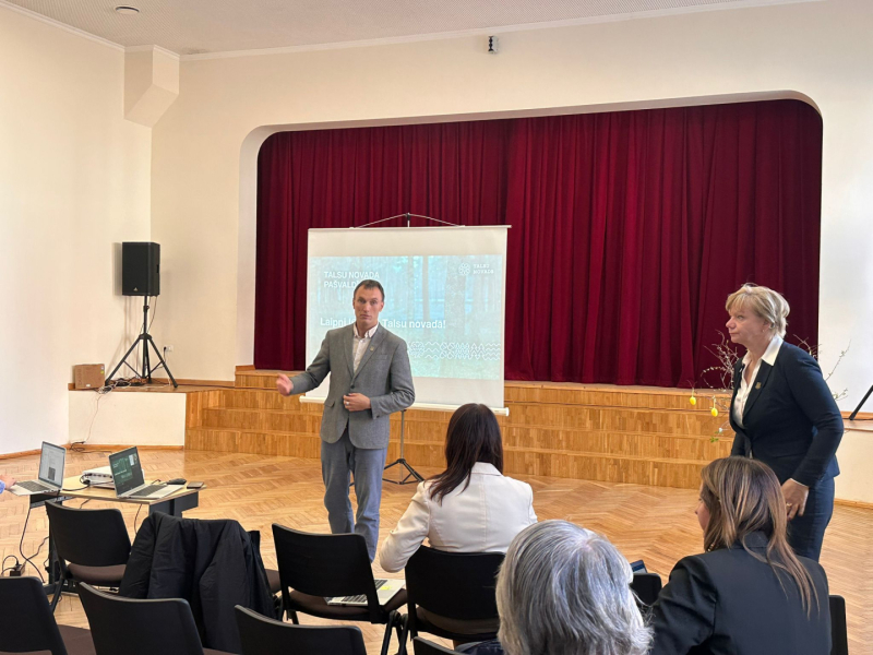 Laucienes kultūras namā Talsu novada pašvaldības tikšanās laikā priekšplānā redzami divi pašvaldības pārstāvji, kuri uzrunā sanāksmes apmeklētājus. Fonā uz ekrāna redzama prezentācija ar uzrakstu „Laipni lūdzam Talsu novadā!”, bet aiz tās – skatuve ar tumši sarkaniem aizkariem. Priekšplānā sēž iedzīvotāji un redzami atvērti portatīvie datori.