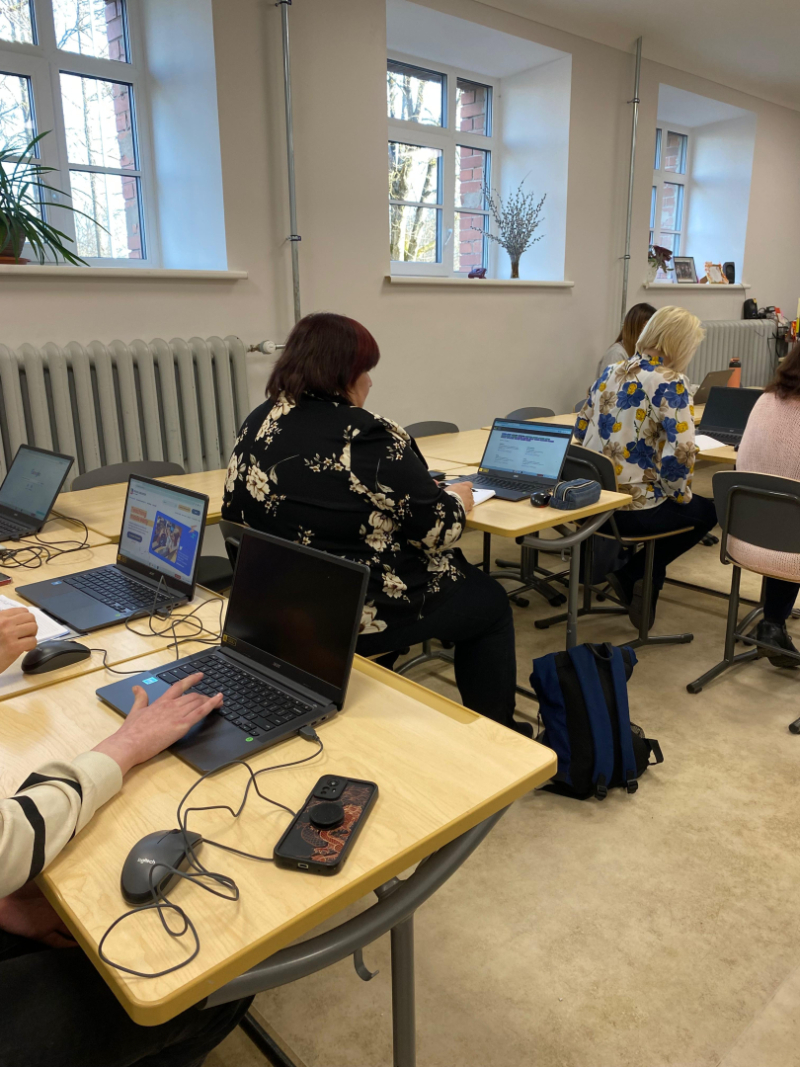 Klasē pie galdiem sēž pedagogi ar klēpjdatoriem un piezīmēm, piedaloties darbseminārā. Priekšplānā redzams galds ar datoru, krūzi un darba materiāliem, bet fonā – dalībnieki, kas klausās un strādā pie uzdevumiem.