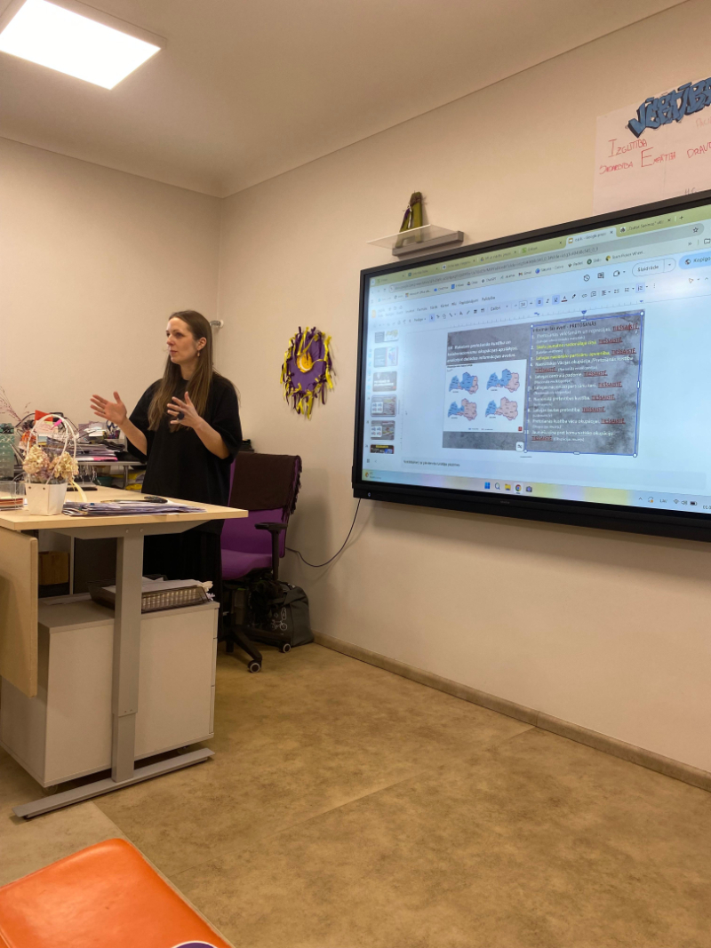Pedagoģe klasē stāv pie interaktīvās tāfeles un vada prezentāciju. Uz ekrāna redzams digitāls mācību materiāls, bet skolotāja ar žestiem skaidro tēmu semināra dalībniekiem.