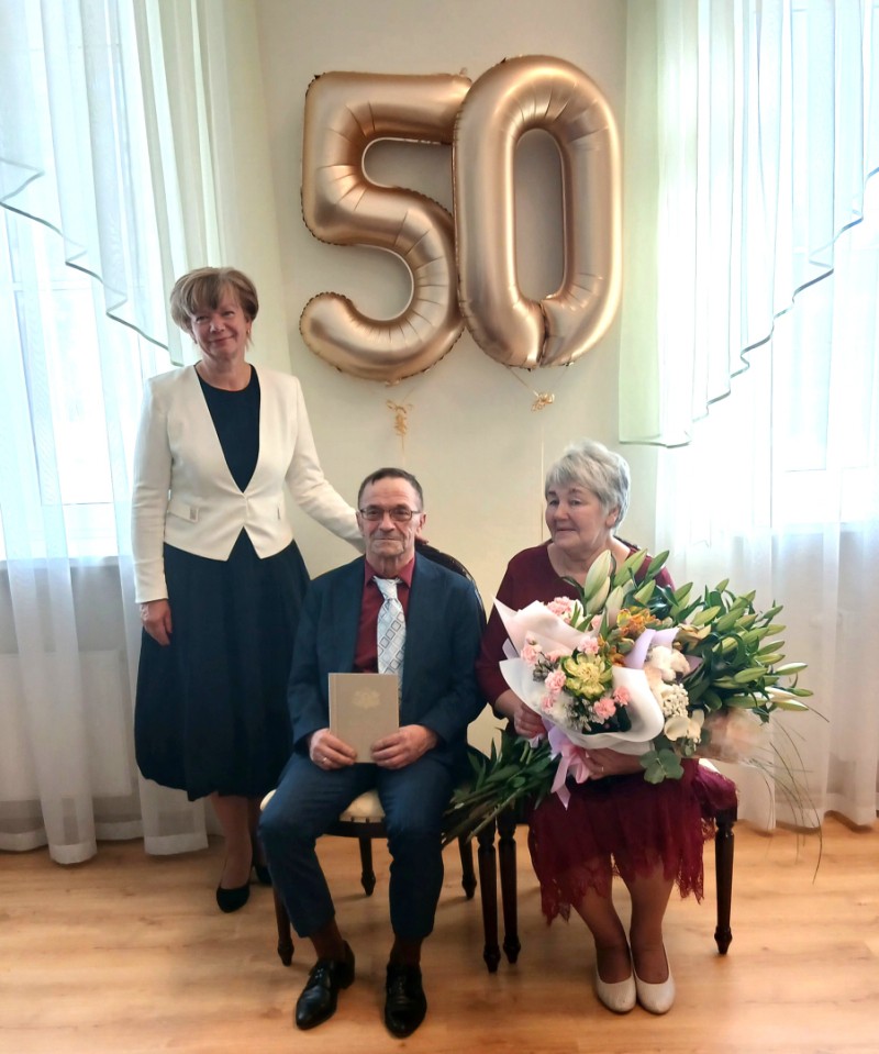 Zelta kāzu jubilāri Ināra un Juris Martinovski svinīgā pasākumā Rojas Kultūras centrā kopā ar Talsu novada domes priekšsēdētāja vietnieci Ligitu Šnori. Fonā redzami zeltaini baloni ar skaitli 50, bet Ināra rokās tur lielu ziedu pušķi.