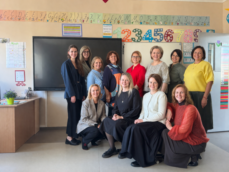 Attēlā klasē redzama pedagogu grupa, kas pozē kopīgai fotogrāfijai. Fonā redzama interaktīvā tāfele, mācību materiāli, alfabēts un ciparu plakāti, kas raksturīgi sākumskolas videi.