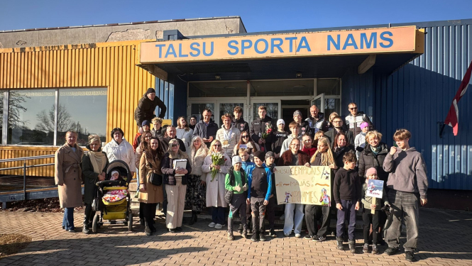 Pie ēkas ieejas redzama cilvēku grupa, kas svinīgi sagaida sportistu, turot rokās apsveikuma plakātu.