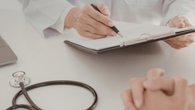 Ārsta kabinetā mediķis baltā halātā raksta piezīmes uz planšetes, kamēr priekšplānā redzamas pacienta sakļautas rokas un uz galda novietots stetoskops.