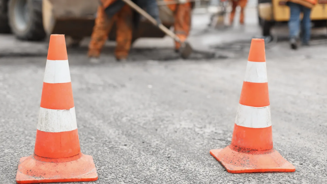 Divi oranži satiksmes konusi uz ceļa seguma priekšplānā, bet fonā izplūduši redzami ceļu būvdarbu strādnieki un tehnika.