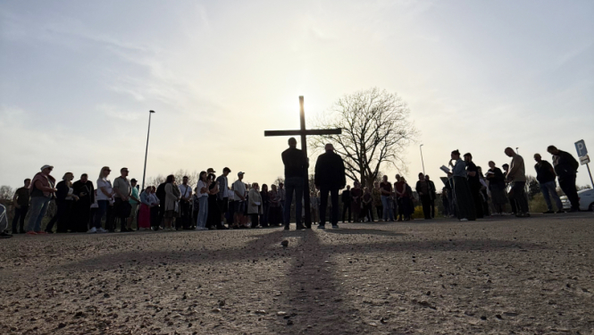 Zema rakursa fotogrāfijā redzams cilvēku loks brīvā dabā ap lielu koka krustu. Priekšplānā redzams grants segums, bet fonā – cilvēku silueti pret saules gaismu, kas atrodas aiz krusta. Pie krusta stāv divi cilvēki, apkārt pulcējušies vairāki desmiti cilvēku. Fonā redzams arī koks un skaidras debesis.
