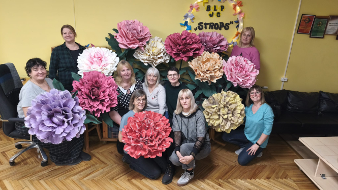 Grupas foto redzamas desmit sievietes Brīvā laika pavadīšanas un jauniešu centra „Strops” telpās, pozējot ar lielformāta papīra ziediem dažādos toņos – rozā, baltā, ceriņkrāsas, sarkanā, krēmkrāsas un dzeltenīgā. Fonā pie sienas redzama centra dekorācija ar uzrakstu „BLP JC “Strops””, bet telpā ir dzeltenas sienas, koka grīda un tumšs dīvāns.
