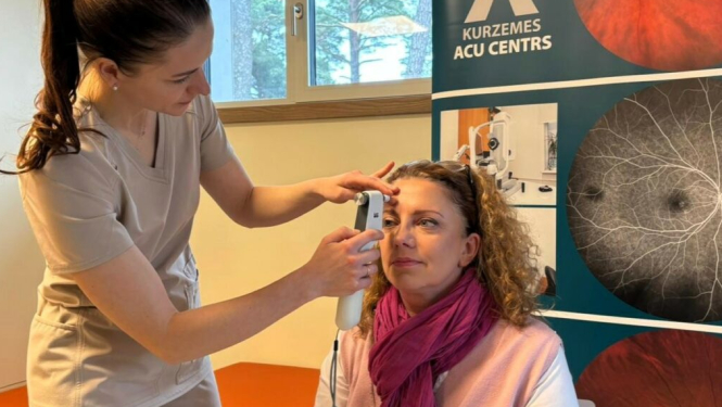 Medicīnas darbiniece veic acu spiediena mērījumu sievietei, kura sēž krēslā, bet fonā redzams “Kurzemes acu centra” stends ar acu izmeklējumu attēliem.