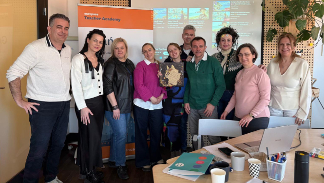 Grupas foto no Erasmus+ mobilitātes Gdaņskā – telpā pie „Europass Teacher Academy” stenda un projekcijas ekrāna pozē vairāki kursu dalībnieki. Priekšplānā redzams galds ar klēpjdatoru, krūzēm, rakstāmpiederumiem un mācību materiāliem, bet daļa dalībnieku rokās tur dekoratīvu darbu.