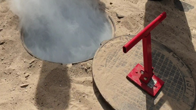 Atvērta kanalizācijas akas lūka smilšainā teritorijā; no akas paceļas balti dūmi vai tvaiks, bet blakus novietots noņemts vāks ar sarkanu rokturi.