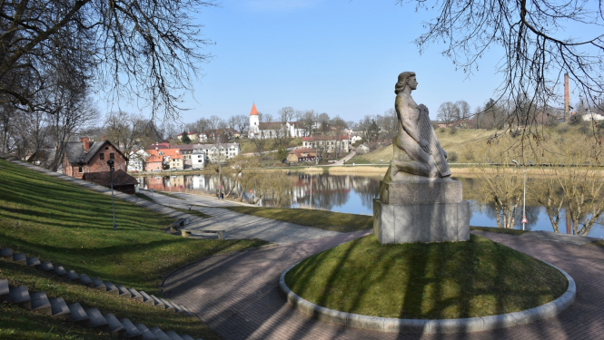 Piemineklis „Koklētājs” Talsos pavasarīgā dienā pie Talsu ezera. Priekšplānā redzama uz postamenta novietota skulptūra, apkārt tai bruģēts celiņš un zāliens, bet fonā ezers, koki bez lapām un Talsu pilsētas apbūve ar baznīcu kalnā.