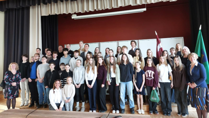 Plašā zālē uz nelielas skatuves kopīgai fotogrāfijai pozē liela jauniešu un pieaugušo grupa. Fonā redzama tumši sarkana siena, balta tāfeļu rinda, Latvijas karogs un zaļš karogs, virs skatuves – melnbalti aizkari un koka griesti ar gaismekļiem. Priekšplānā rindās izvietojušies skolēni un pieaugušie, daļa stāv, daļa sēž vai pietupjas. Attēls rada svinīgas un draudzīgas kopbildes iespaidu, visticamāk pēc kāda pasākuma vai tikšanās skolā vai kultūras centrā.