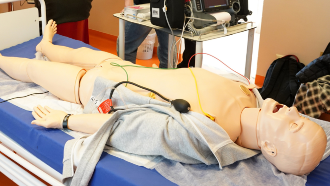 Mācību telpā uz gultas novietots medicīnas simulators ar pieslēgtiem vadiem un monitorēšanas aprīkojumu, kamēr vairāki cilvēki fonā vēro informāciju uz ekrāna.