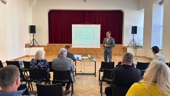 : Laucienes kultūras namā notiek Talsu novada pašvaldības tikšanās ar iedzīvotājiem. Zāles priekšā pie ekrāna stāv runātājs, bet klausītāji sēž krēslu rindās un seko prezentācijai. Fonā redzama skatuve ar tumši sarkaniem aizkariem, skaļruņi abās pusēs un galds ar projektoru un portatīvajiem datoriem.