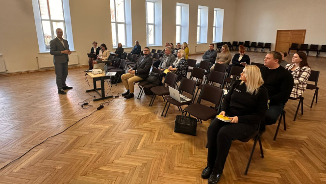 Laucienes kultūras nama zālē notiek Talsu novada pašvaldības tikšanās ar iedzīvotājiem. Priekšplānā redzams runātājs pie projektora galdiņa, bet zālē rindās sēž sanāksmes apmeklētāji un pašvaldības pārstāvji. Plašajā telpā ir gaišas sienas, augsti logi un koka grīda.