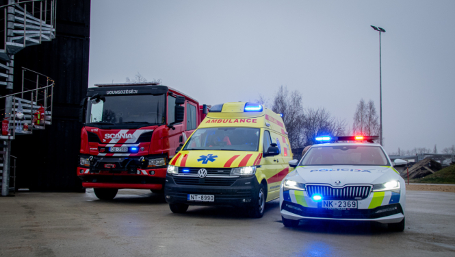 Attēlā redzami trīs operatīvie transportlīdzekļi – ugunsdzēsēju automašīna, Neatliekamās medicīniskās palīdzības dienesta automašīna un Valsts policijas dienesta auto – novietoti blakus uz laukuma. Visām automašīnām ieslēgtas bākugunis. Fonā redzama ēka, laterna un koki apmākušās debesīs.