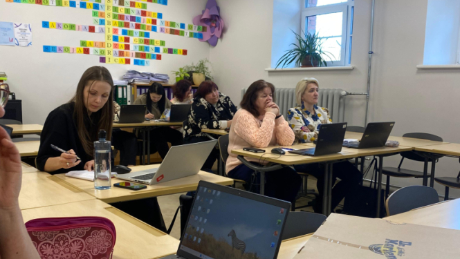 Klasē pie galdiem sēž pedagogi ar klēpjdatoriem un piezīmēm, piedaloties darbseminārā. Priekšplānā redzams galds ar datoru, krūzi un darba materiāliem, bet fonā – dalībnieki, kas klausās un strādā pie uzdevumiem.