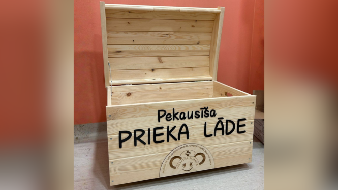 Atvērta koka kaste ar uzrakstu „Pekausīša prieka lāde”, novietota uz galda pie oranžas sienas. Uz kastes redzams arī Pekausīša slimnīcas logo.