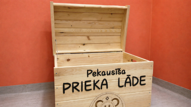Atvērta koka kaste ar uzrakstu „Pekausīša prieka lāde”, novietota uz galda pie oranžas sienas. Uz kastes redzams arī Pekausīša slimnīcas logo.