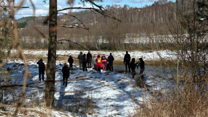 Cilvēku grupa agrā pavasara dienā stāv sniegainā upes vai dīķa krastā, fonā redzami kaili koki un zilas debesis ar mākoņiem. Skats fotografēts no neliela uzkalna caur koku zariem, priekšplānā redzama iemīta taka.