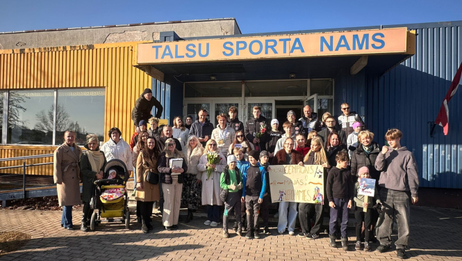 Pie ēkas ieejas redzama cilvēku grupa, kas svinīgi sagaida sportistu, turot rokās apsveikuma plakātu.
