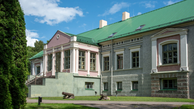 Attēlā redzama Talsu novada muzeja galvenā ēka – plaša, vēsturiska celtne ar zaļu metāla jumtu, gaiši pelēku un rozā fasādi, lieliem logiem un dekoratīviem arhitektūras elementiem. Ēkas priekšā redzams zāliens, celiņš un vairāki vēsturiski lielgabali, bet fonā – koki un zilas debesis ar mākoņiem.