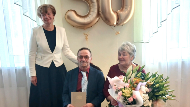 Zelta kāzu jubilāri Ināra un Juris Martinovski svinīgā pasākumā Rojas Kultūras centrā kopā ar Talsu novada domes priekšsēdētāja vietnieci Ligitu Šnori. Fonā redzami zeltaini baloni ar skaitli 50, bet Ināra rokās tur lielu ziedu pušķi.