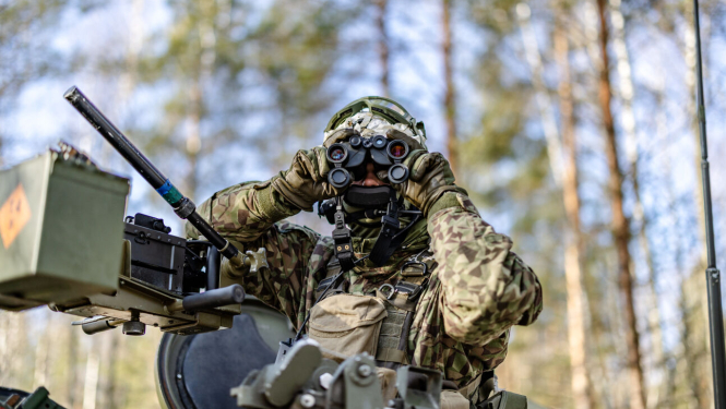 Mežainā apvidū redzams karavīrs maskēšanās formā un ekipējumā, kurš caur binokli vēro apkārtni. Viņš atrodas pie militārās tehnikas aprīkojuma, bet fonā redzami koki un skaidras debesis.