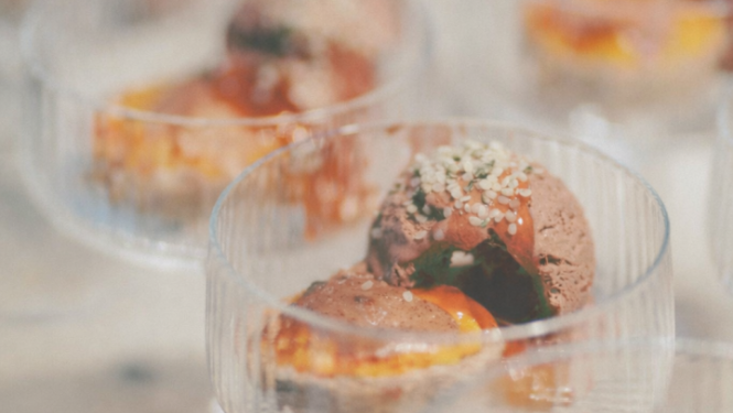 Tuvplāna fotogrāfijā redzami vairāki stikla trauciņi ar desertu – šokolādes saldējuma bumbiņām un oranžīgu mērci. Priekšplānā fokusā ir viens deserts ar sēkliņu pārkaisījumu, bet pārējie trauciņi fonā izplūduši maigā, siltā gaismā.