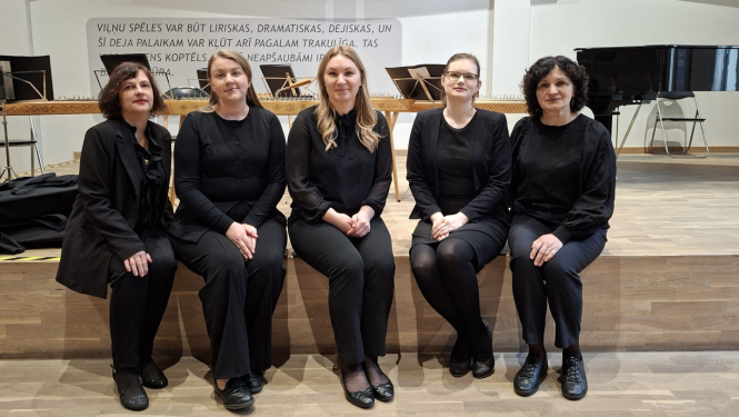 Attēlā redzamas piecas sievietes melnā koncerttērpā, sēžot uz skatuves malas. Fonā redzamas kokles, nošu pultis un flīģelis, kas liecina par muzikālu uzstāšanos vai koncertu.