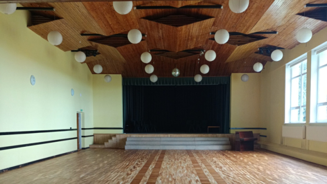 Lubes kultūras nama zāle – plaša koka grīda, koka griesti ar apaļiem gaismekļiem, skatuve ar aizkariem un logi gar sānu sienu.