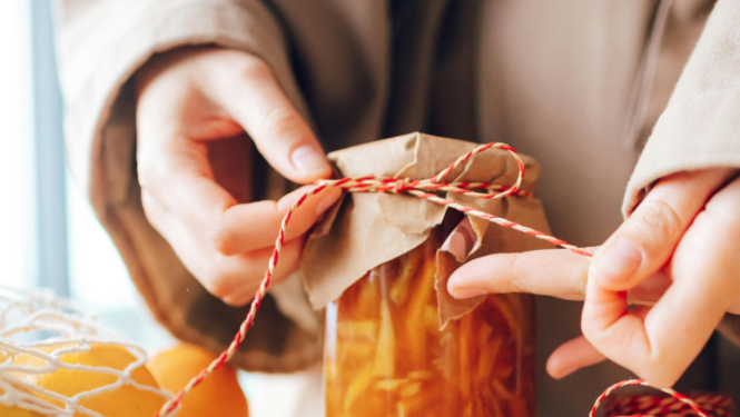 Tuvplānā redzamas rokas, kas apsien aukliņu ap stikla burciņu ar ievārījumu vai sīrupu. Burciņa pārklāta ar brūnu papīru, bet blakus uz galda atrodas citrusaugļi, koka karote un sarkani balta aukla, radot siltu, mājīgu noskaņu.