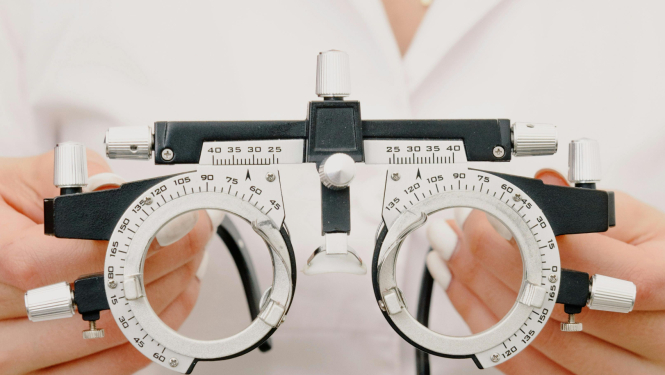 Optometrists tur rokās redzes pārbaudes ierīci (foropteru) ar regulēšanas skrūvēm un graduētām skalām, gatavojoties noteikt briļļu lēcu stiprumu.