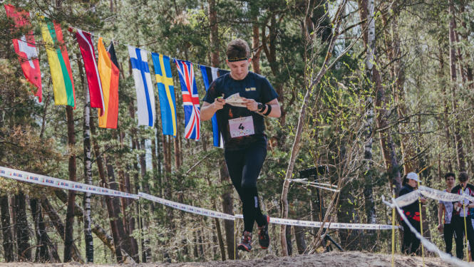 Orientierists skrien pa smilšainu meža posmu, lasot karti; fonā starp kokiem izkārti dažādu valstu karodziņi un redzami sacensību norobežojumi.