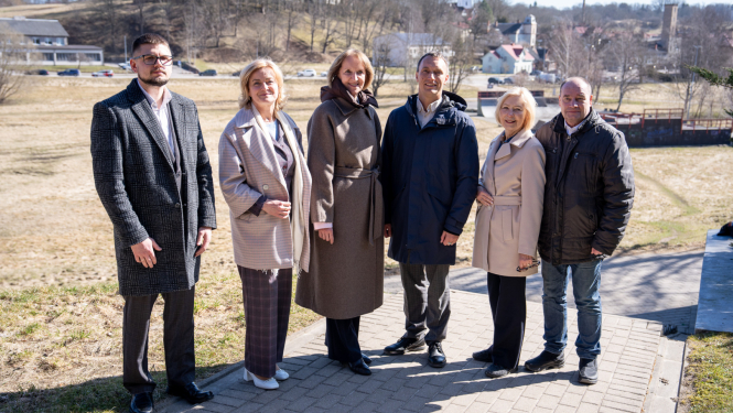 Alt teksts:  Pieci cilvēki un viens jaunietis stāv ārā uz bruģēta celiņa saulainā pavasara dienā. Fonā redzama Talsu ainava ar pakalniem, kokiem un pilsētas ēkām, tostarp baznīcas torņiem. Grupa pozē kopīgai fotogrāfijai, visi ģērbušies mēteļos un virsjakās.