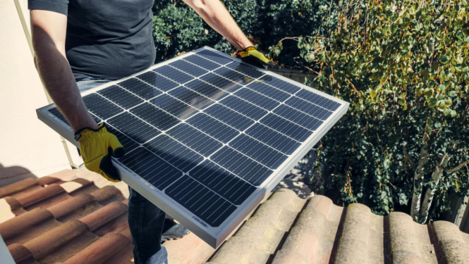 Attēlā redzams cilvēks, kurš uz ēkas jumta tur rokās saules paneli, gatavojoties tā uzstādīšanai. Fonā redzami koki un saulains laiks.