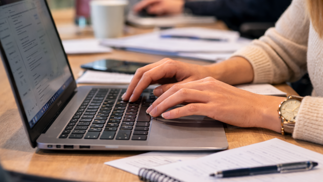 Tuvplāna foto redzamas rokas, kas raksta uz klēpjdatora tastatūras pie darba galda; blakus novietota piezīmju klade un pildspalva, fonā izplūdis biroja darba process.