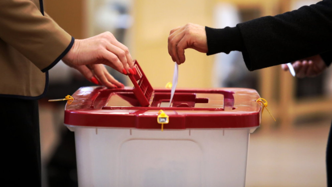 Tuvplānā redzama balsošana vēlēšanu iecirknī – divi cilvēki vienlaikus ievieto vēlēšanu zīmes plastmasas balsošanas kastē ar sarkanu vāku. Uz kastes redzamas drošības plombas, bet fonā izplūdusi iekštelpu vide.