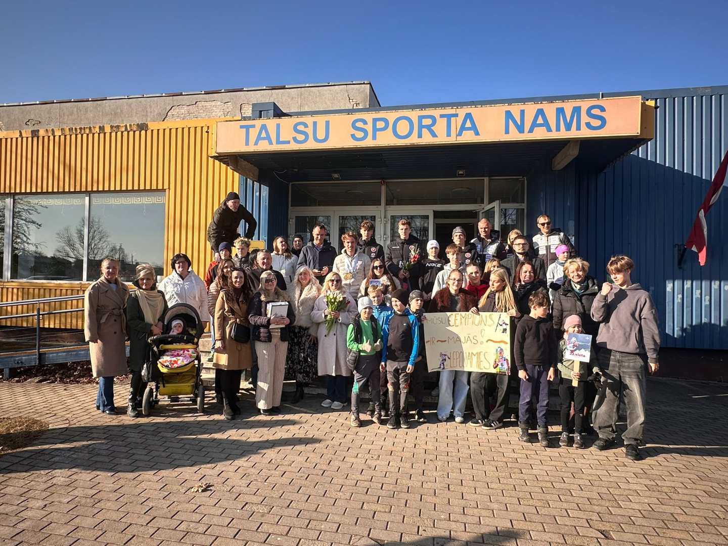 Pie ēkas ieejas redzama cilvēku grupa, kas svinīgi sagaida sportistu, turot rokās apsveikuma plakātu.