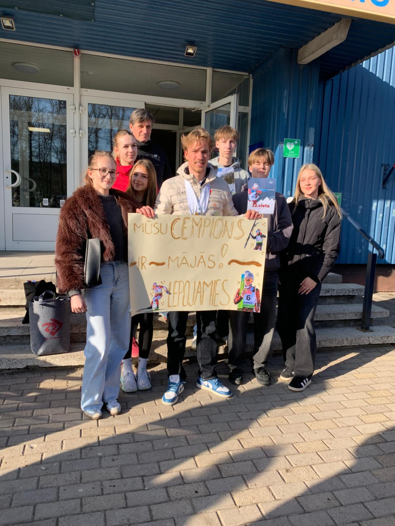 Pie ēkas ieejas redzama cilvēku grupa, kas svinīgi sagaida sportistu, turot rokās apsveikuma plakātu.