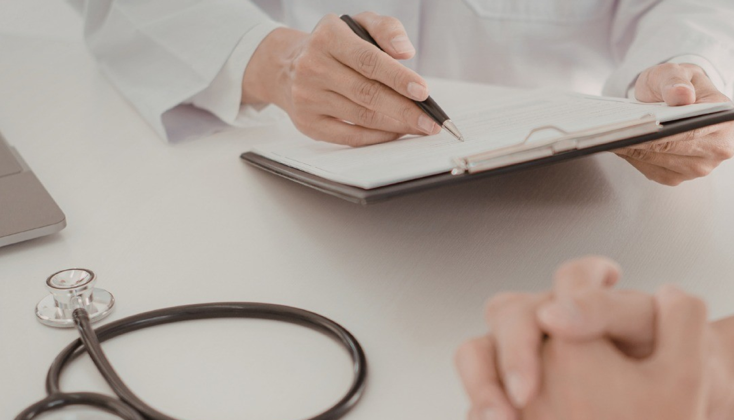 Ārsta kabinetā mediķis baltā halātā raksta piezīmes uz planšetes, kamēr priekšplānā redzamas pacienta sakļautas rokas un uz galda novietots stetoskops.