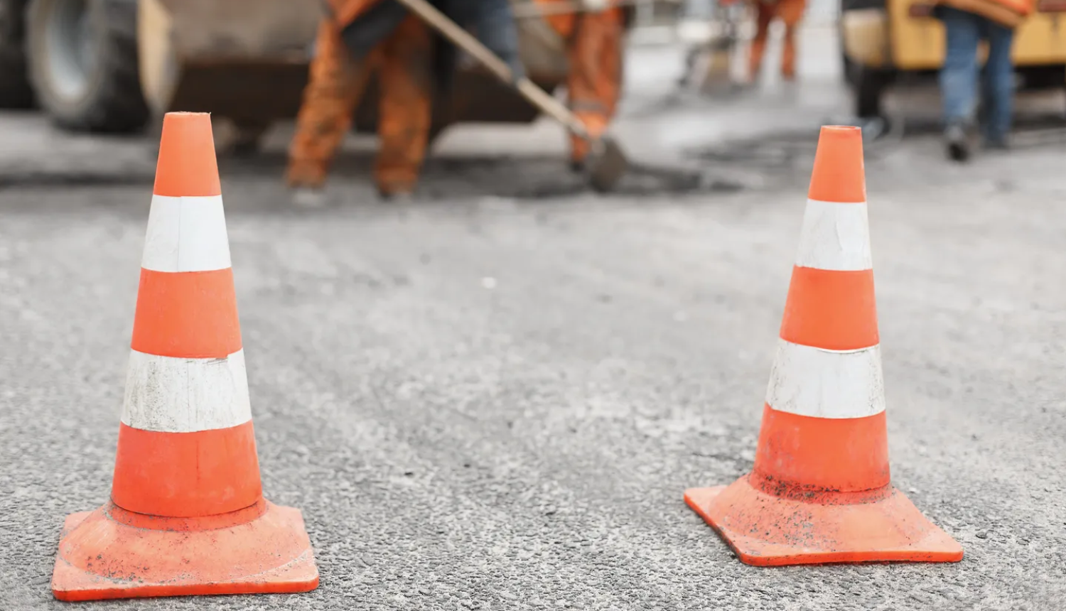 Divi oranži satiksmes konusi uz ceļa seguma priekšplānā, bet fonā izplūduši redzami ceļu būvdarbu strādnieki un tehnika.